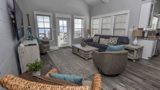 A living room with a sofa, coffee table, and a TV. There are two windows with blinds, a kitchen area, and a staircase leading to another level. - Rising Sun Pool Elevator  Pet Beach Access (Eastpoint)