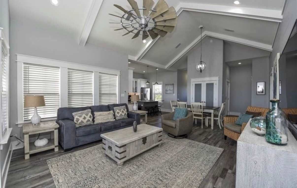 A spacious living area with a large ceiling fan, a sofa set, a dining table, and a kitchen in the background.