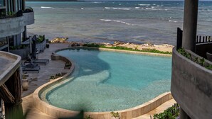Une piscine à bordures courbes, entourée d’une terrasse en béton et de plantes en pot, offrant une vue sur une plage et l’océan.