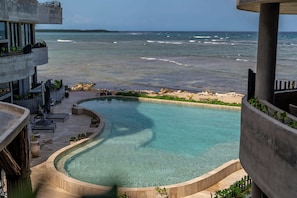 A pool with a curved edge, surrounded by a concrete deck and potted plants, overlooking a beach and ocean. - Oceanfront luxury 3BR in Tankah w/ Private Beach (Tulum)