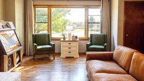 Una habitación con suelo de madera, un sofá de cuero marrón, dos sillones verdes, un aparador blanco y una ventana con cortinas.