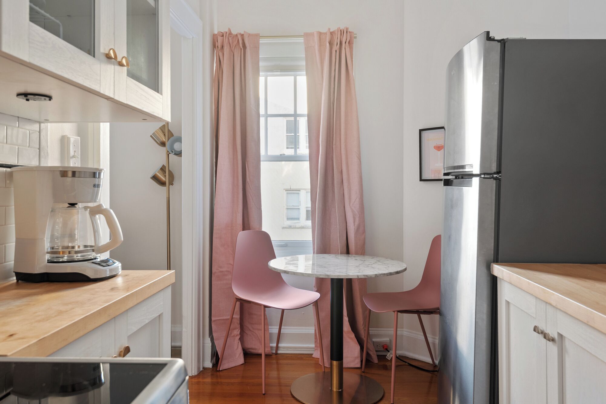 A compact kitchen with a coffee maker, two pink chairs, a refrigerator, and a window with curtains.