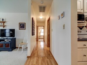 A hallway with a wooden floor, a television, a white chair, and kitchen cabinets.