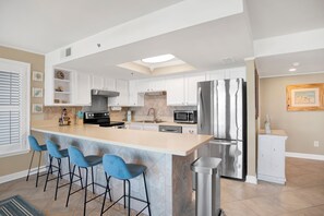A modern kitchen with white cabinets, a stainless steel refrigerator, a microwave, and a bar area with blue bar stools. - 3-bedroom Ocean front condo in Ocean Beach. Skip the elevator, keep the view! (Orange Beach)