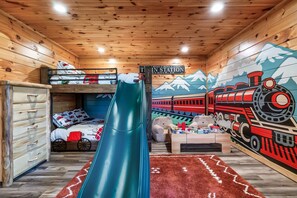 A cabin interior with a bunk bed, a slide, a play area with toys, and a mural of a train. - Hot Tub Hideaway! 5 Min to Parkway | Cozy n Quiet! (Pigeon Forge)