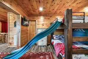 L’intérieur d’une cabane en bois, avec un lit superposé, un toboggan bleu, un jeu de fléchettes et un tableau accroché au mur.