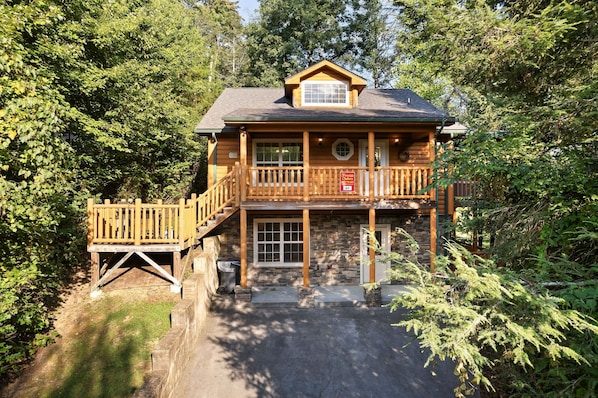 A two-story log cabin with a balcony, a stone foundation, and a wooden deck. - Hot Tub Hideaway! 5 Min to Parkway | Cozy n Quiet! (Pigeon Forge)