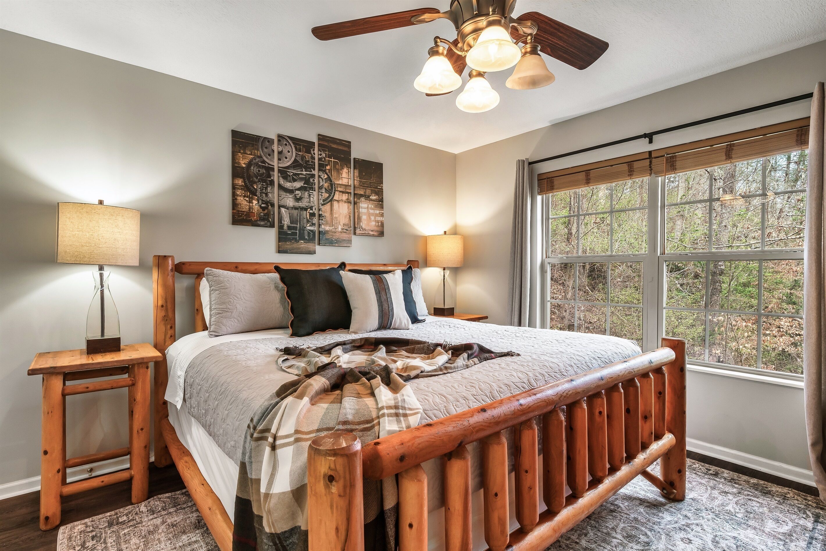 A bedroom with a wooden bed frame, a large window with a view of trees, and a ceiling fan with lights.