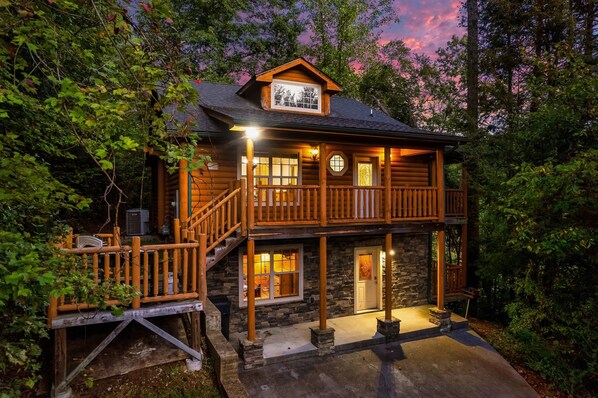 A two-story log cabin with a balcony, stone foundation, and a porch with a railing. - Hot Tub Hideaway! 5 Min to Parkway | Cozy n Quiet! (Pigeon Forge)