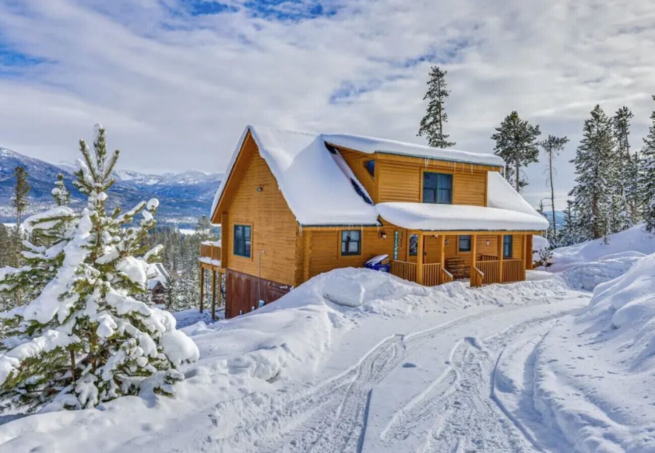A wooden cabin with a sloped roof, surrounded by snow-covered trees and mountains.