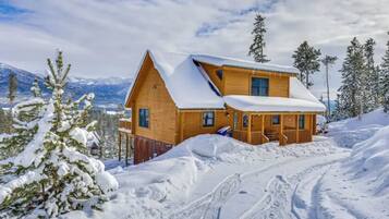 A wooden cabin with a sloped roof, surrounded by snow-covered trees and mountains.