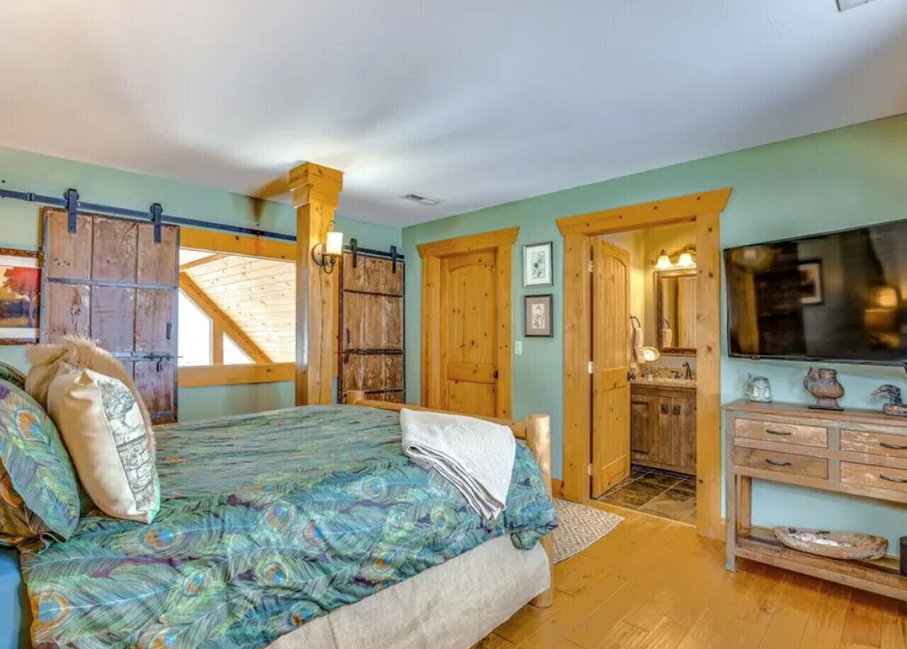 A bedroom with a bed, wooden walls, a door leading to a kitchen, and a large window.