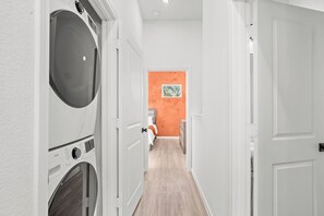 Une buanderie équipée d’un lave-linge et d’un sèche-linge, un couloir menant à une chambre dont un mur est peint dans une teinte orange, et une porte donnant sur une autre pièce.