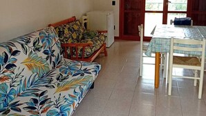 Una sala de estar con un sofá, una mesa de comedor con sillas, un refrigerador y una puerta que da al exterior.