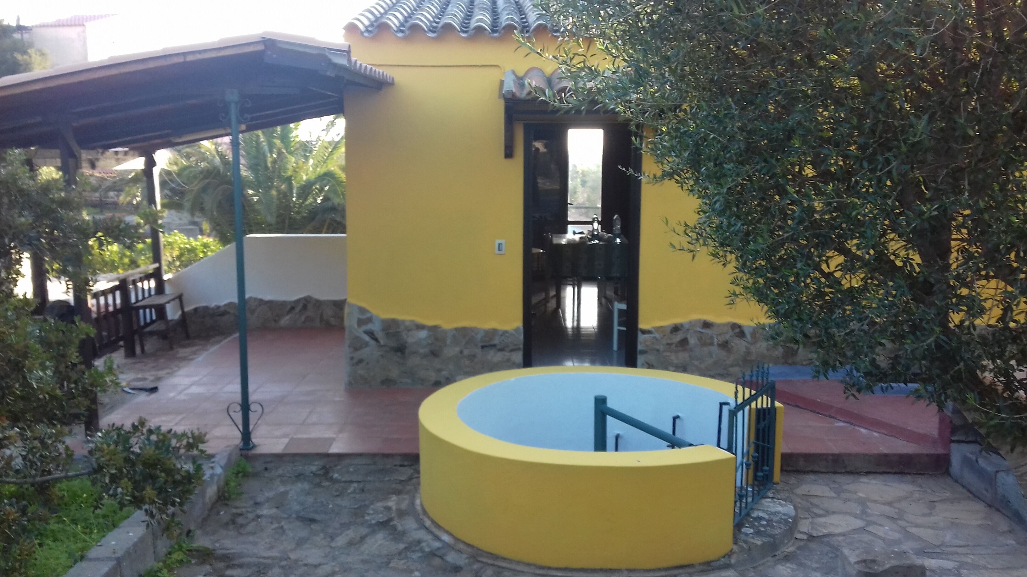 Um edifício amarelo com base de pedra, alpendre coberto e piscina circular.
