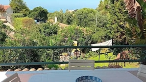 Una mesa blanca con sillas en una terraza, con vistas a un jardín exuberante y una vista distante del mar.