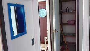 Un pasillo con un espejo de marco azul, una puerta blanca y una habitación con una cama, una silla y una estantería con objetos.