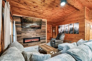 Ein gemütliches Blockhaus-Interieur mit Kamin, einer bequemen Couch, einem Ledersessel und einer Holzbalkendecke.