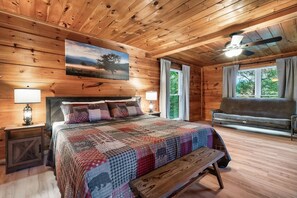A wooden cabin bedroom with a bed, a bench, a sofa, a painting, and a ceiling fan. - Falcon's Rest | 5 Min to Gatlinburg | Sleeps 18 (Gatlinburg)