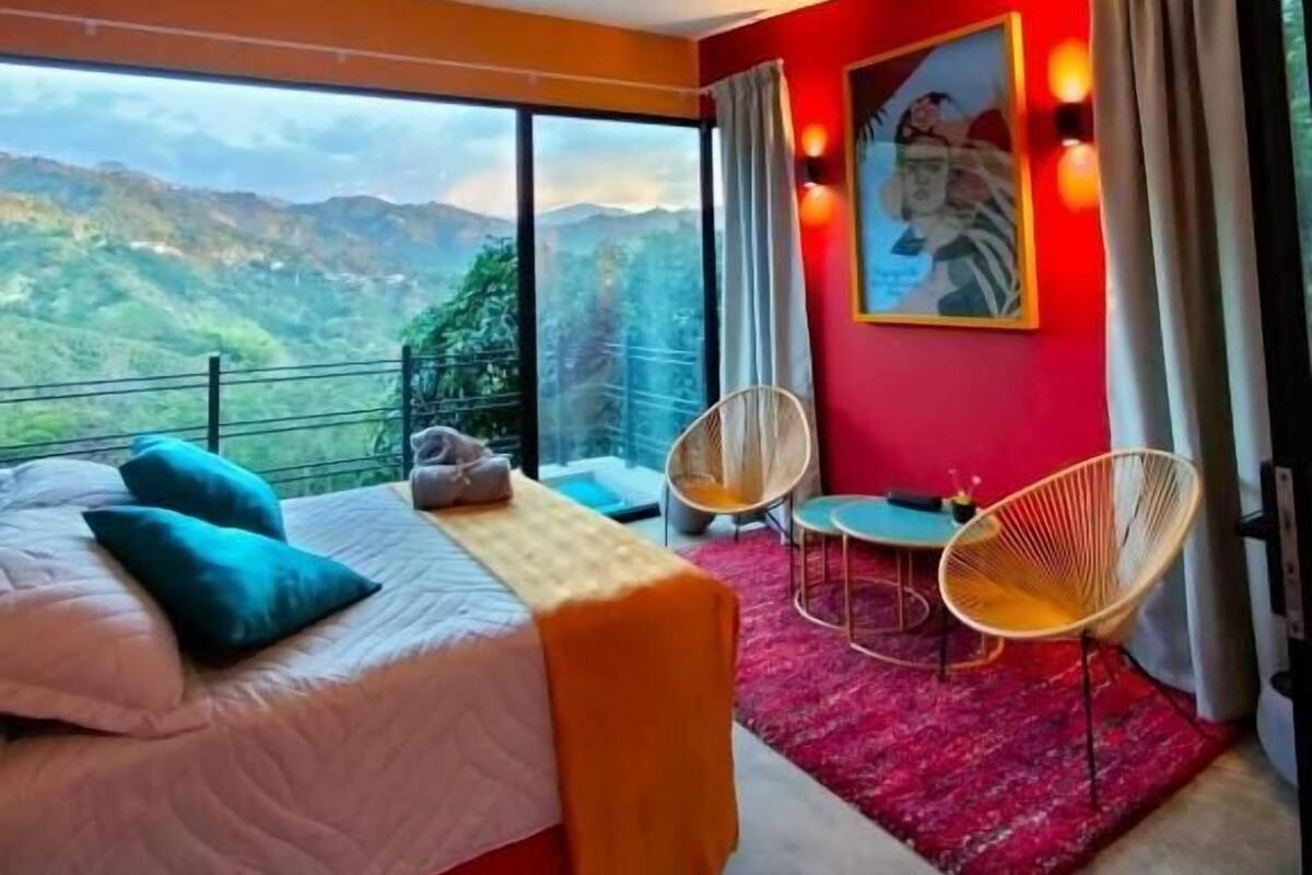 Un dormitorio con cama, dos sillones, una mesita y un ventanal con vistas a las montañas.