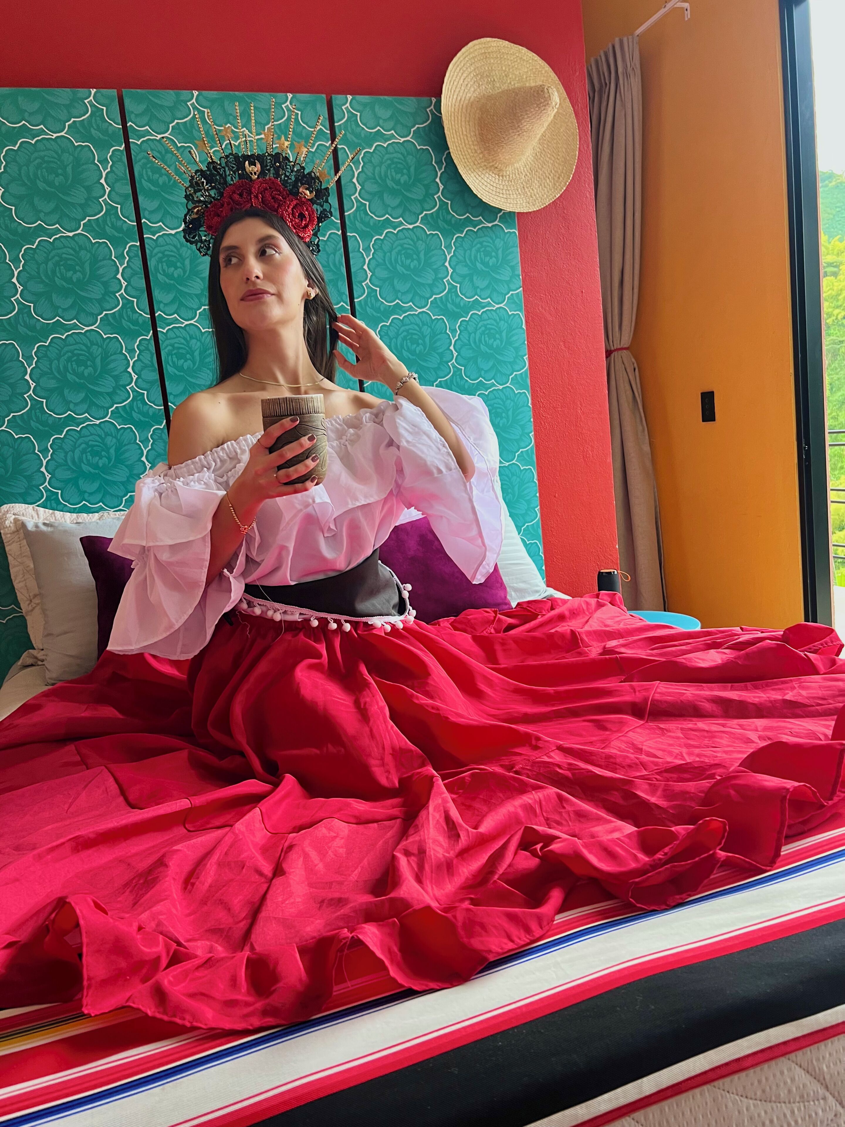 A person sitting on a bed with a red and white striped bedspread, a teal patterned wall, and a straw hat on a hanger.