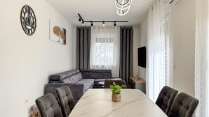 A modern living room with a sectional sofa, a dining table with chairs, a potted plant, and a wall-mounted clock. - Cozy home in Nedescina (Nedescina)
