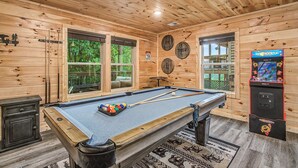 A pool table with a pool cue and balls, a claw machine, and wooden walls with windows. - The Brookshire! Premium View | Hot Tub | Top 10% (Sevierville)