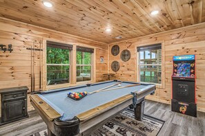 A pool table with a pool cue and balls, a claw machine, and wooden walls with windows. - The Brookshire! Premium View | Hot Tub | Top 10% (Sevierville)