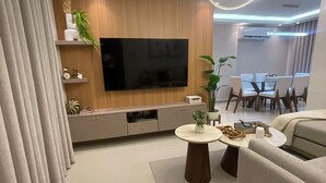 A modern living room with a flat-screen TV, a sofa, a dining table, and a ceiling light fixture.