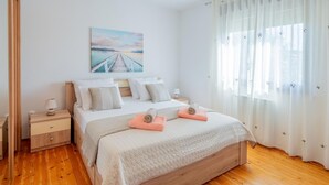 Een slaapkamer met een houten bed, wit beddengoed, een nachtkastje, een lamp, een schilderij van een brug en dunne gordijnen.