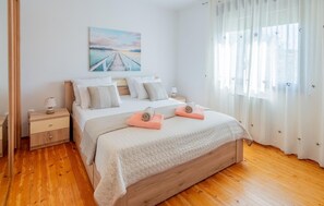 A bedroom with a wooden bed, white bedding, a nightstand, a lamp, a painting of a bridge, and sheer curtains.