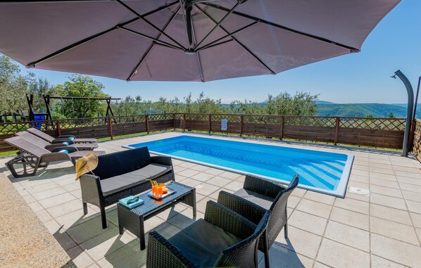 A swimming pool with lounge chairs and an umbrella, surrounded by a wooden fence and trees.