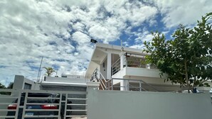 A gated entrance with a car parked inside, a white building with a balcony, and a tree to the right. - Lovely apartment with AC in vibrant Punta Cana. 10min to Beach & Downtown. (Punta Cana)