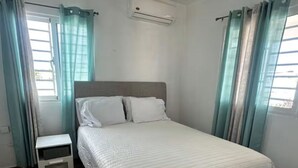 A bedroom with a bed, a nightstand, an air conditioning unit, and curtains. - Lovely apartment with AC in vibrant Punta Cana. 10min to Beach & Downtown. (Punta Cana)