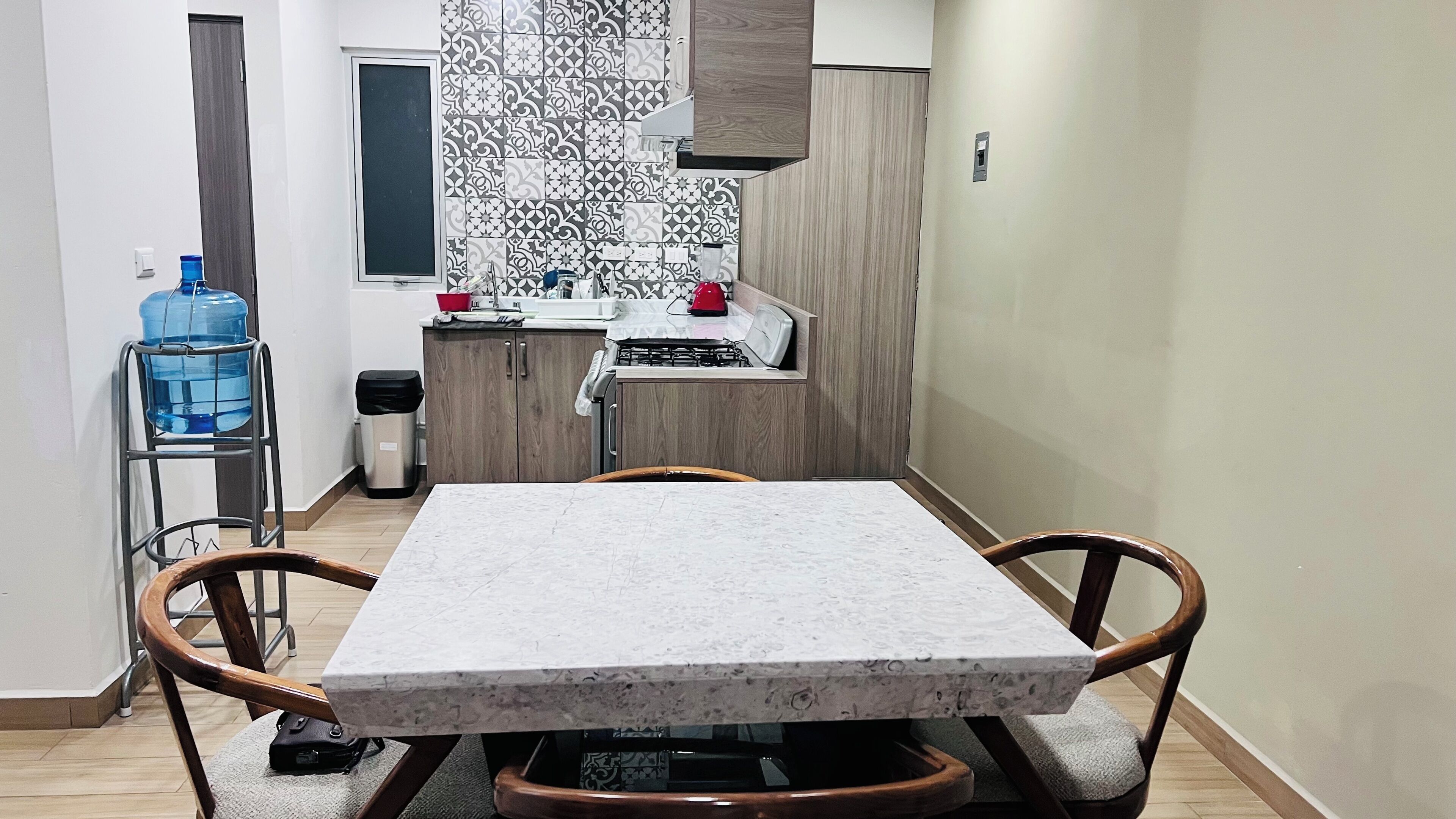 A kitchen with a marble countertop, wooden cabinets, a water dispenser, and a trash can.