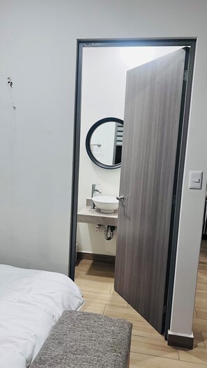 Une chambre à coucher comprenant un lit, une table de chevet et une porte donnant sur une salle de bain avec un lavabo et un miroir.
