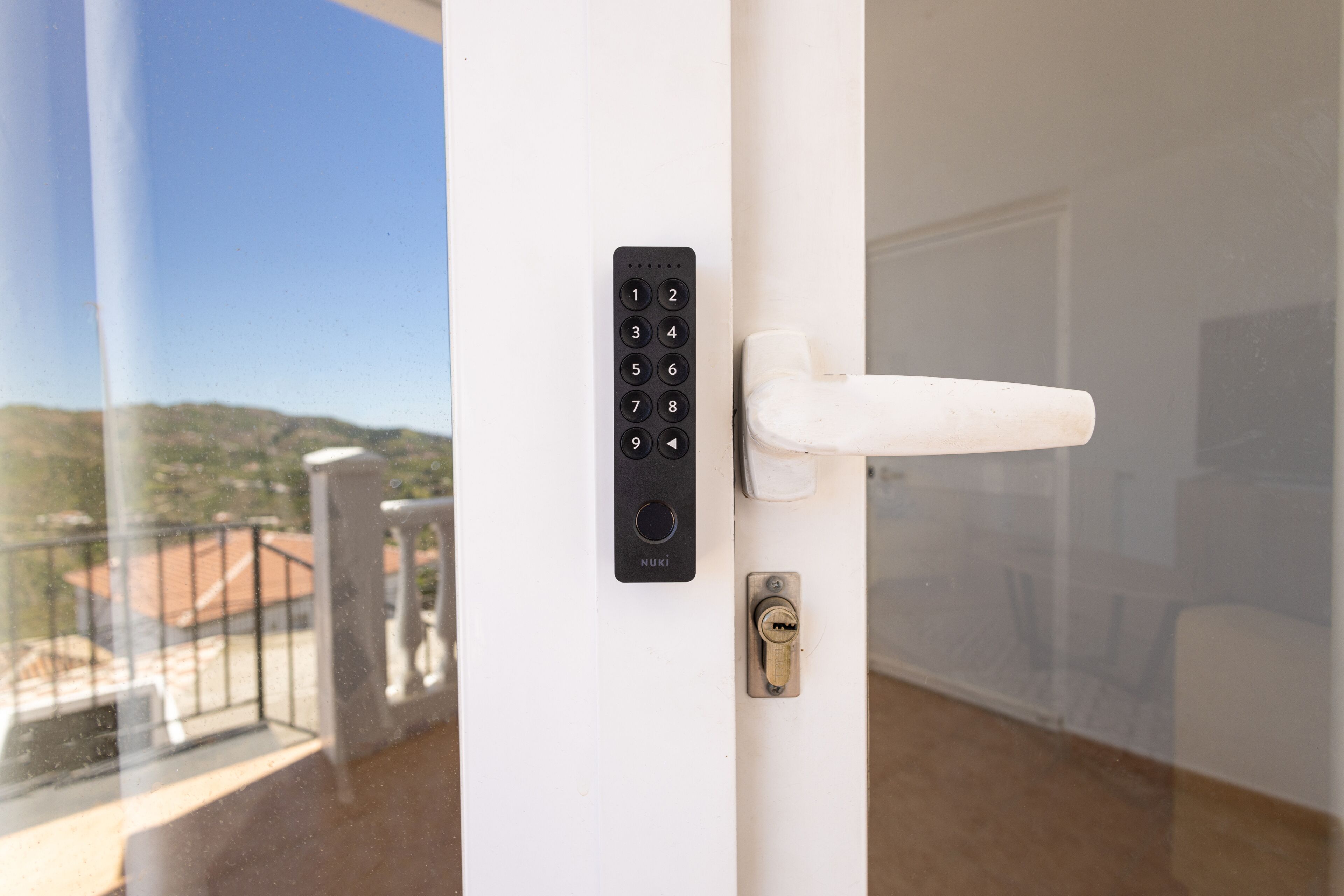 A door with a key lock and a keypad, a balcony with a railing, and a view of a building with a red roof.