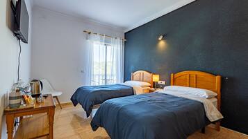 A hotel room with two beds, a TV mounted on the wall, a desk with a lamp, and a window with curtains.