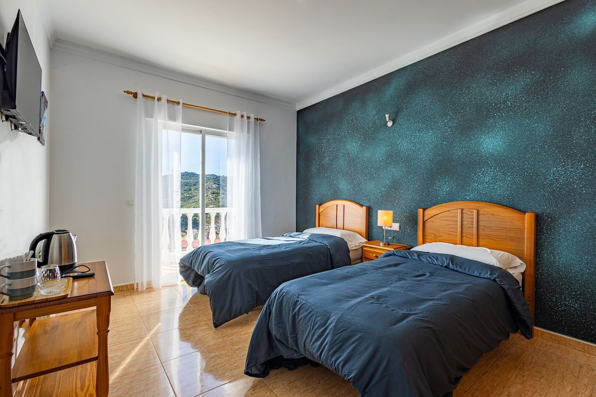 Ett hotellrum med två sängar, ett träbord med en vattenkokare, utsikt över omgivningarna och en vägg med en intressant yta.