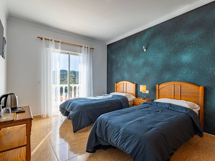 Une chambre d’hôtel avec deux lits, une table en bois avec une bouilloire, une vue sur l’extérieur et un mur à texture.