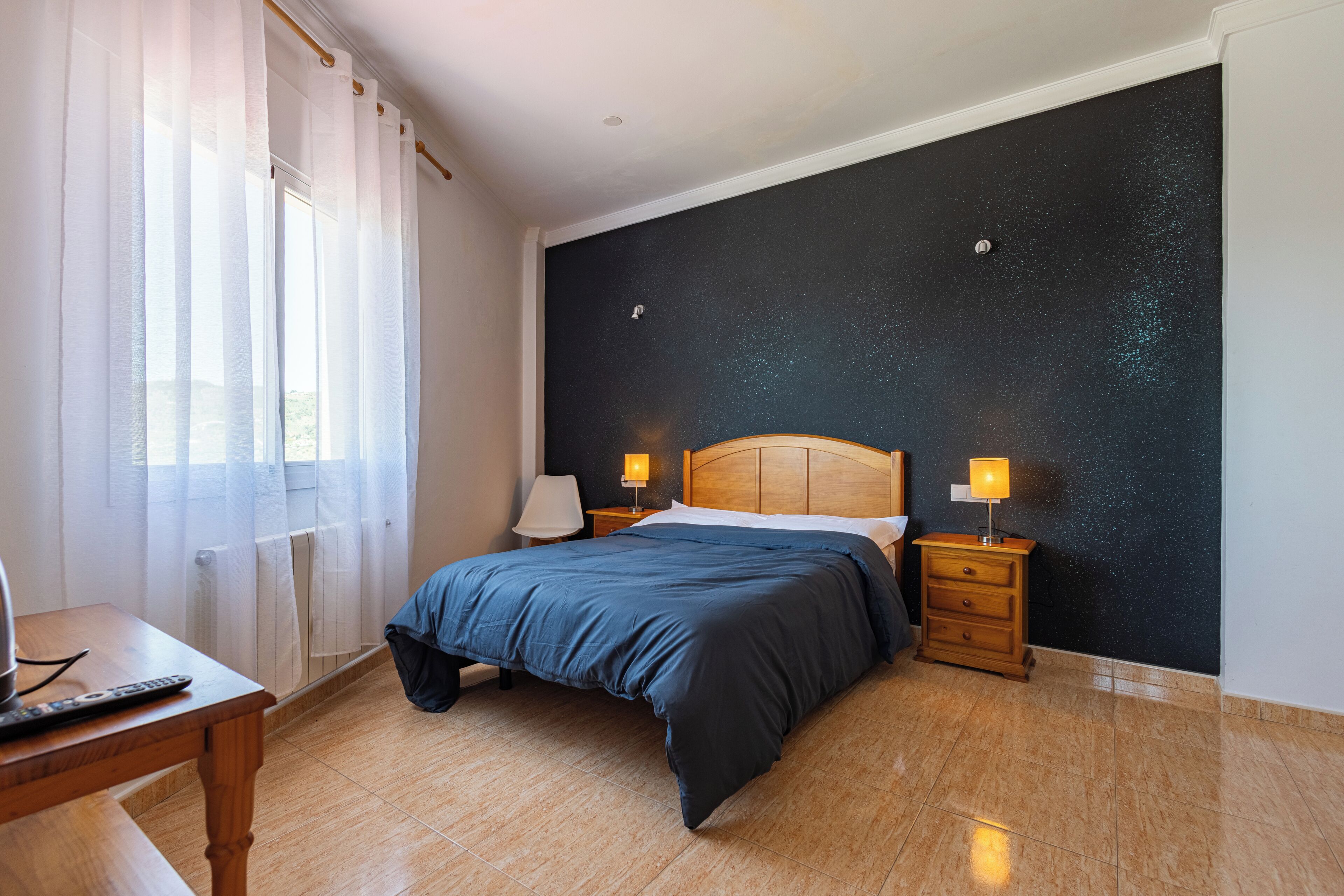 A bedroom with a bed, bedside tables, a lamp, a window with curtains, and a wall with a starry pattern.