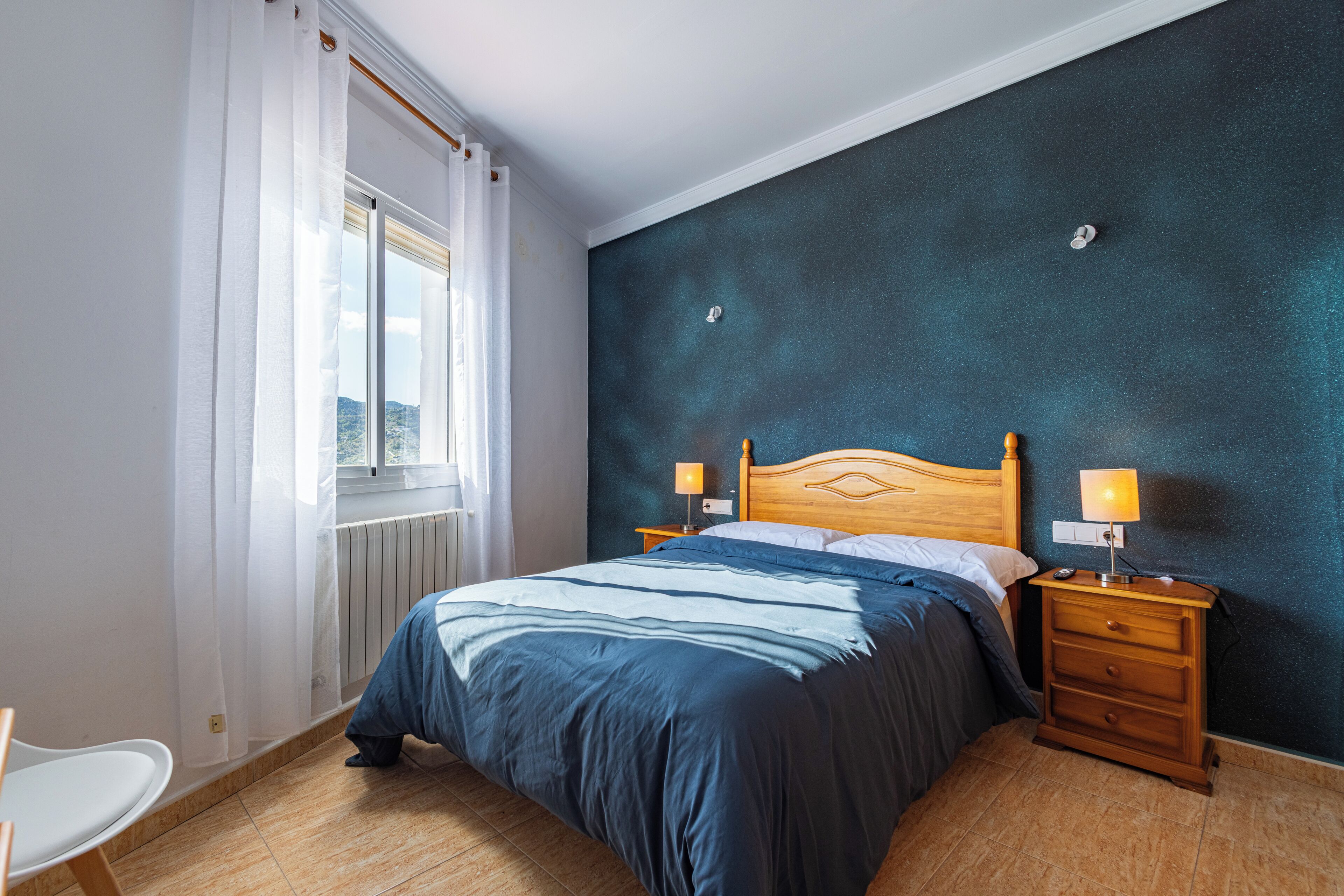 A bedroom with a bed, two bedside tables with lamps, a window with curtains, and a radiator.