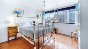 Un dormitorio con estructura de cama metálica, una mesita de noche, una silla, un ventanal con vista a la ciudad y una araña.