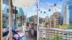 Un balcón con sillas y una mesa, con vistas a un paisaje urbano con edificios y zonas verdes.