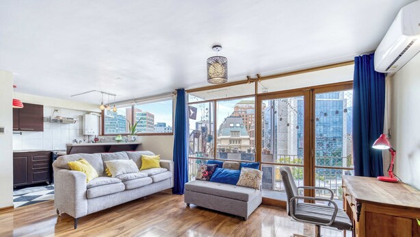 Una sala moderna con un sofá gris, un pequeño reposapiés, un escritorio de madera y una silla. Amplias ventanas ofrecen vistas a la ciudad.