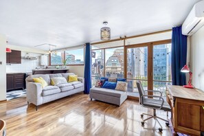 Una sala moderna con un sofá gris, un pequeño reposapiés, un escritorio de madera y una silla. Amplias ventanas ofrecen vistas a la ciudad.