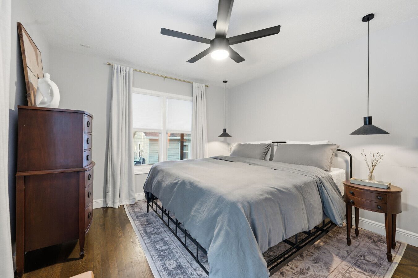 Un dormitorio con cama, ventilador de techo, mesita de noche, cómoda y ventana con cortinas.
