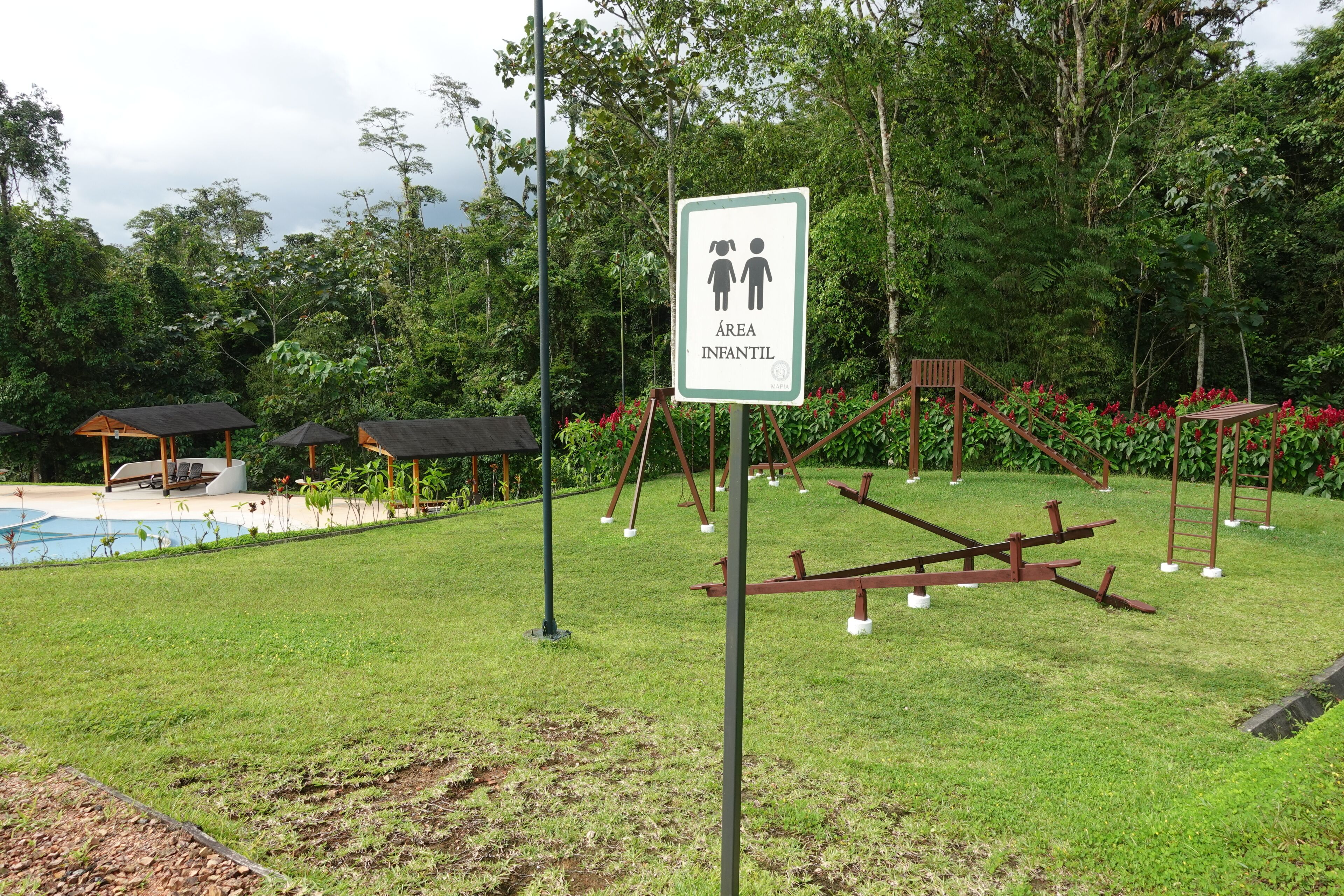 Un panneau indiquant un espace de jeux pour enfants, une piscine et des structures de jeux en bois, le tout dans un cadre verdoyant et luxuriant.