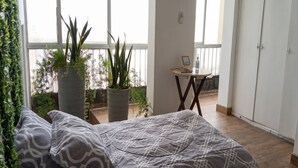 A bedroom with a bed, a small table with a picture frame, and potted plants near a window. - Historic Center Quito by Otium (Quito)