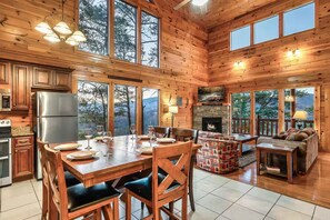 Interior de una cabaña de madera con comedor, cocina, chimenea y ventanas amplias.